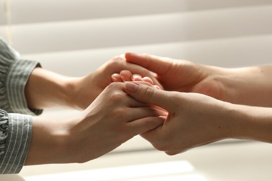 Religious women holding hands and praying together near window indoors, closeup