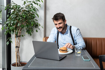 Young freelancer business man on lunch break eating hamburger in cafeteria and surfing the net on his laptop. Successful business marketing expert learning on online course while he eating. E-comerce