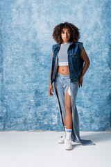 Wall Mural - full length of trendy african american woman in torn jeans and denim vest standing on blue background