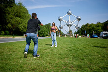 A Young Hispanic Male Taking A Photo Of His Girlfriend Posing At Atomium, Brussels, Belgium