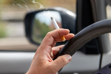 Close-up detail view of male hand driver holding cigarette while driving inside car on higway city street road on day time. Man person smoking inside vehicle. Low concentration attention danger