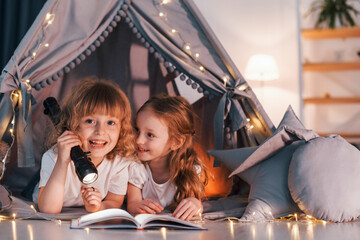 Reading book and using flashlight. Two little girls is in the tent in domestic room together