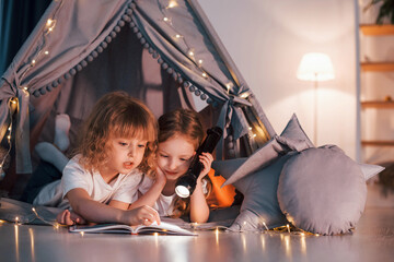 Wall Mural - Laying down and using flashlight to read the book. Two little girls is in the tent in domestic room together