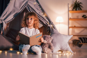 Wall Mural - Cute little girl with curly hair is in the tent in domestic room with interesting book