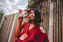 Portrait Of Sexy And Young Japanese Girl With Beautiful Old Traditional Red Kimono And Handmade Cat Mask In Front Of A Bamboo Wall In A Cloudy Day