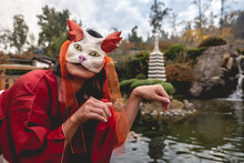 Portrait Of Sexy And Young Japanese Girl With Beautiful Old Traditional Red Kimono And Handmade Cat Mask In Front Of A Pond In A Japanese Garden