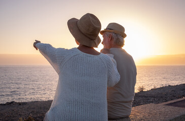 Sticker - Back view of two relaxed and romantic seniors or pensioners sitting face to the sea at sunset light looking at the horizon - old couple outdoors enjoying vacations together