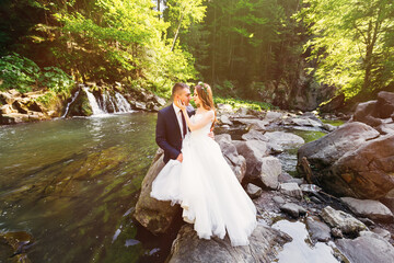 Beautiful newlyweds sit on stones and hug. Stylish wedding recep