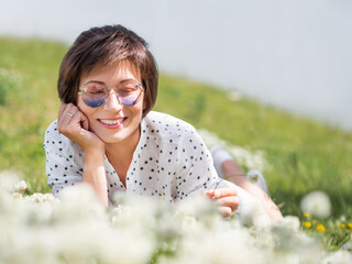 Wall Mural - Woman in colorful sunglasses sniffs clover flowers on lawn in urban park. Nature in town. Relax outdoors after work. Summer vibes.