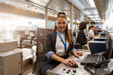 Woman Airline Employee Working At Airline Check In Counter In Airport With Colleagues On Background
