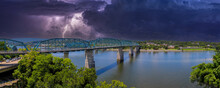 A Panoramic Shot Of The Rippling Waters Of The Tennessee River With The Walnut Street Bridge Over The Water Surrounded By Lush Green Trees With Powerful Storm Clouds And Lightning In Chattanooga