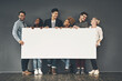 A copyspace of a diverse group of young, happy and smiling professional business people holding a blank white billboard. A multiracial team of men and women against a dark wall with copy space