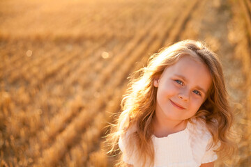 Adorable little curly girl 4 years old in a white dress in the sun at sunset in a mowed field of wheat. Happy child outside. Walk. Warm summer. Emotions.