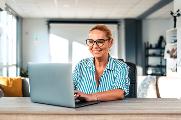 Smiling middle age woman sitting at the table at office working using computer laptop with a happy face standing and smiling with a confident smile showing teeth.