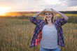 © olga_sova - A beautiful middle-aged farmer woman in a straw hat and a plaid shirt stands in a field of golden ripening wheat during the daytime in the sunlight