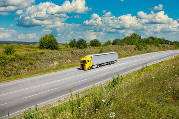 Wall Mural - yellow cargo truck on the highway. asphalt road among green fields and beautiful clouds. cargo delivery and transportation concept