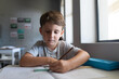 Caucasian elementary schoolboy writing on book while sitting at desk in classroom