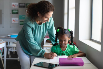 Wall Mural - Caucasian young female teacher assisting african american elementary schoolgirl writing on book