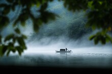 View Of Silhouette Of Man In Small Fishing Boat Reflected On Surface Of Water On Thick Foggy Lake
