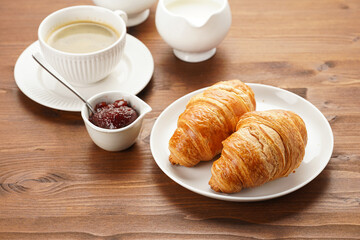 Wall Mural - Two french soft croissants on a white plate on a wooden table, a cup of black coffee in white sophisticated cup, milk, sugar, jam