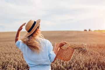 Wall Mural - Back view of young stylish woman in hat walking in summer field holding straw handbag with wheat bundle.