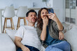 © wifesun - Cheerful in love couple taking a selfie on the phone while sitting on the couch together, making funny mustaches out of their hair and taking pictures