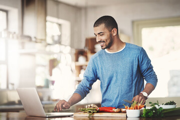 Wall Mural - Young casual man in the kitchen on a laptop while preparing a healthy meal at home. Happy smiling male browsing and learning on computer on how to cook. Guy alone checking online recipes on the web.