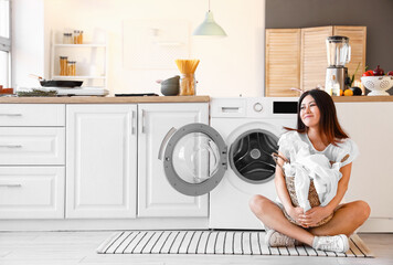 Wall Mural - Young Asian woman with laundry basket sitting near washing machine in kitchen