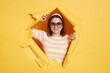 © sementsova321 - Portrait of attractive positive woman wearing hair band and striped t shirt standing in yellow paper hole, looking at camera with toothy smile, being in good mood.