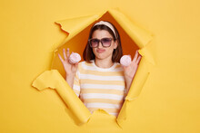Portrait Of Attractive Woman Wearing Hair Band And Striped T Shirt Standing In Yellow Paper Hole, Holding Marshmallow And Frowning Face, Being On Diet, Looking At Camera.