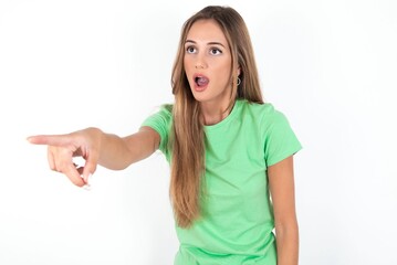 Wall Mural - young beautiful woman wearing green T-shirt over white background Pointing with finger surprised ahead, open mouth amazed expression, something on the front.