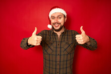 Smiling Man In Christmas Hat, Casual Clothes Shows Thumbs Up With Both Hands Isolated On Red Studio Background With Space For Text. Concept - Holidays, New Year, Promotion, Discounts, Favorable Offer