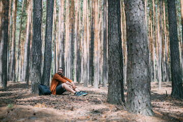 Portrait of a young man sitting at the foot of a pine tree in a coniferous forest on a sunny day. A guy in a shirt is resting under a tree