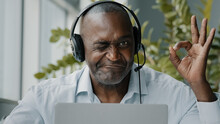 Concentrated African American Senior Entrepreneur Businessman Freelance Worker Wears Headphones With Microphone Chatting Online Browsing Internet Webinar Using Computer App Show Okay Recommend Gesture