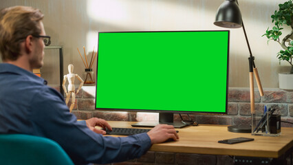 Wall Mural - Young Handsome Man Working from Home on Desktop Computer with Green Screen Mock Up Display. Creative Male Checking Social Media, Browsing Internet. Living Room in Bright Loft Apartment.