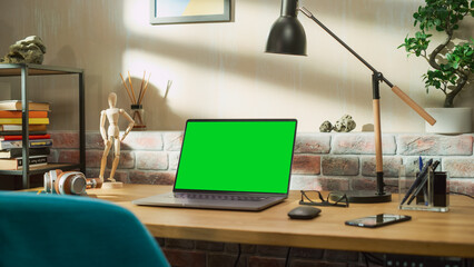 Poster - Laptop Computer Standing on a Wooden Desk with a Green Screen Chromakey Mock Up Display. Cozy Empty Loft Apartment with a Lamp, Notebooks, Smartphone and Headphones on the Table.