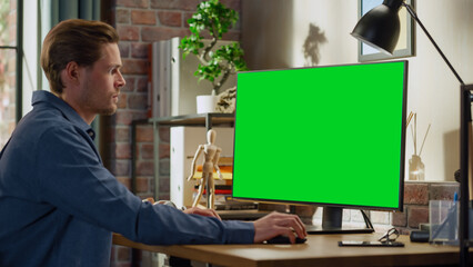 Fototapete - Young Handsome Man Working from Home on Desktop Computer with Green Screen Mock Up Display. Male Checking Corporate Accounts, Messaging Colleagues. Loft Living Room with Big Window.