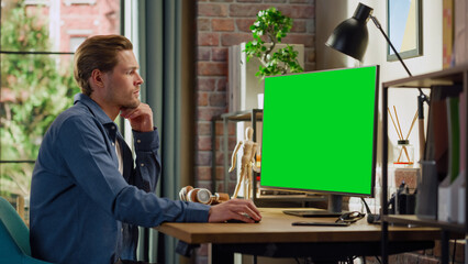 Fototapete - Young Handsome Man Working from Home on Desktop Computer with Green Screen Mock Up Display. Male Checking Corporate Accounts, Messaging Colleagues. Loft Living Room with Big Window.