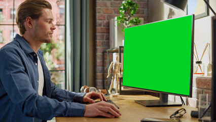 Leinwandbilder - Young Handsome Man Working from Home on Desktop Computer with Green Screen Mock Up Display. Male Checking Corporate Accounts, Messaging Colleagues. Loft Living Room with Big Window.
