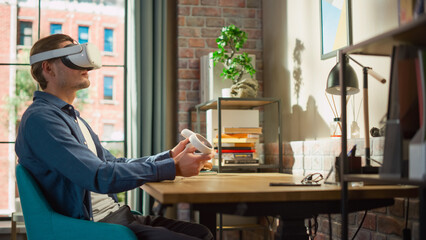 Fototapete - Portrait of a Man Sitting at a Desk at Home or Creative Agency, Using a Virtual Reality Headset and Controllers. Young Male Working from Living Room, Experiencing Futuristic Social Media Concepts.