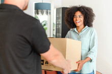 Back View Of Courier Giving Cardboard Box To Happy African American Woman. Cheerful Multiracial Girl With Natural Afro Hairstyle Receiving A Package At Home From A Delivery Guy, Ordered Online