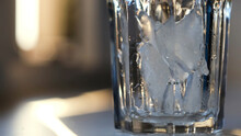 Throwing Ice Pieces Into A Transparent Glass, Preparing Cocktail. Concept. Close Up Of Crystal Glass Standing On The Table Filled With Cold Ice On Blurred Background.
