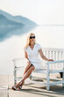 middle aged sexy smiling caucasian woman with sunglasses sits on sea shore in white dress.