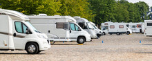 Many White Modern Campervan Recreational Motor Home Vehicles Parked In Row At Camper Park Site Magdeburg City Against Elbe River Bridge. Motorhome Campground Stataion Travel Destination. RV Lifestyle