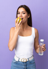 Poster - Young caucasian woman isolated on purple background with a bottle of water and eating an apple