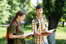 Portrait Of Students In A City Park, Teenage Schoolchildren A Boy And A Girl Walk Along A Path Through The Park And Read Books, Education And Back To School Concept
