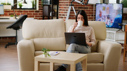 Poster - Smiling young adult freelancer enjoying remote work while sitting on couch inside modern flat. Happy asian woman sipping beverage while working remotely on portable computer at home.