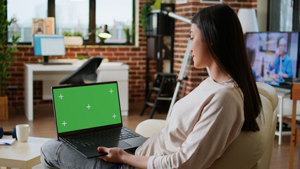 Poster - Young adult person sitting on couch while using laptop having chroma key isolated background. Woman sitting on couch inside apartment with portable computer having green screen display.