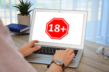 Woman using laptop with age limit sign 18+ years indoors, closeup