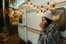 Caucasian Woman In A Knitted Hat Wrapped In A Plaid And Drinks A Warming Drink Outdoors. Travel In A Motor Home In The Fall.